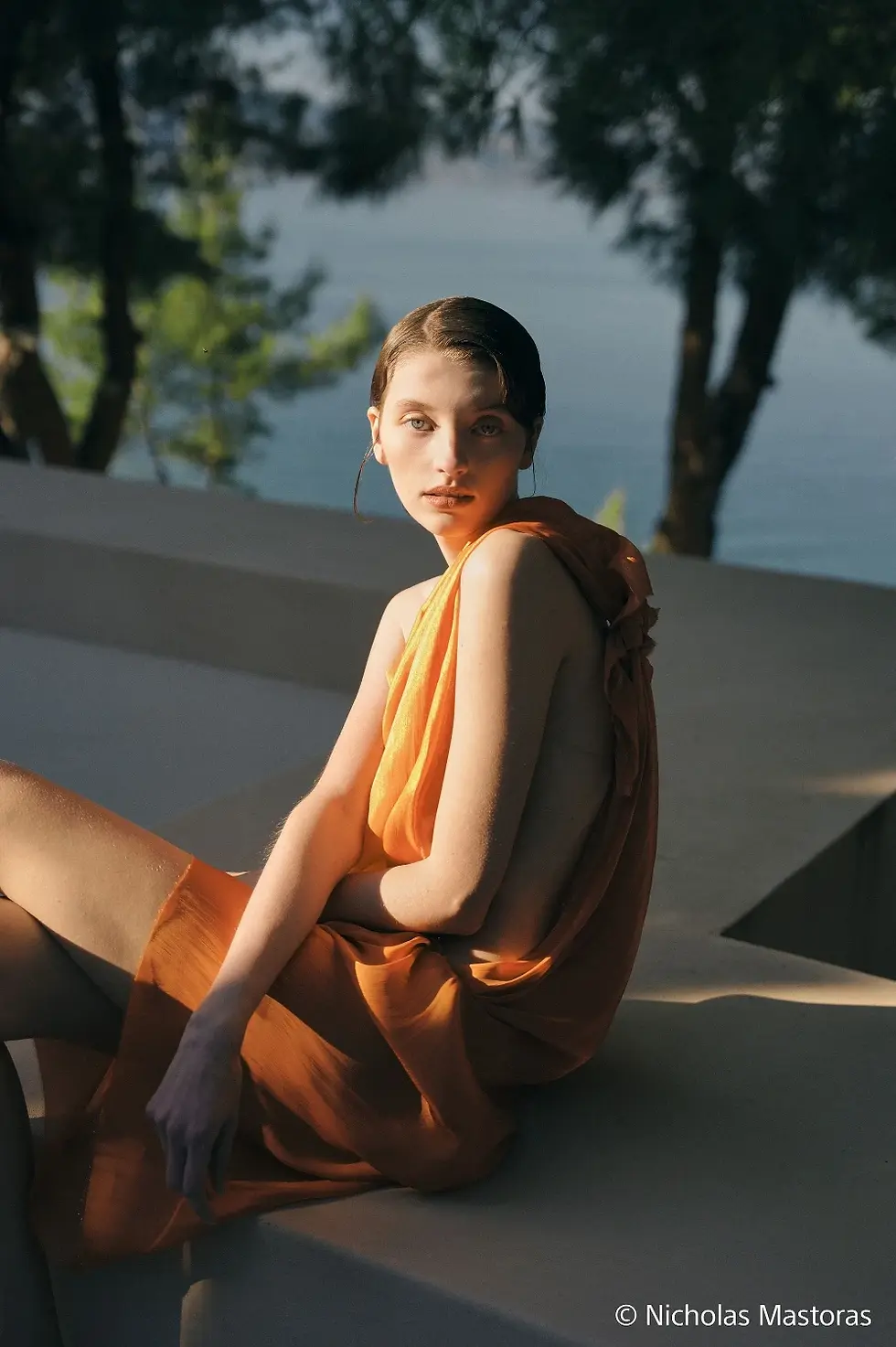 Jeune femme en robe orange assise au bord d’une piscine, regard calme.