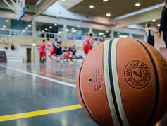 A basketball rests on the court's edge as players compete in an intense indoor game in a well-lit gymnasium.