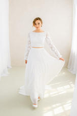 Eine Frau in einem weißen Zweiteiler, im Gang mit Gardinen. lieben.tanzen.lachen. Brautmode Erfurt