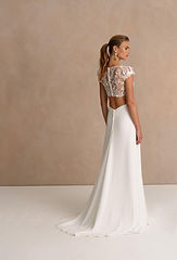 Ein Brautmodel im Brautkleid - La Thuile, steht seitlich und strahlt in einem traumhaften, bodenlangen weißen Kleid mit zartem Sptitzenoberteil und freiem Rücken - ein Moment voller Anmut, Eleganz und stiller Vorfreude.