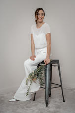 Frau in weißem Zweiteiler, lächelt, sitzt auf Hocker, Blätterspitze im Oberteil und Crepe Hose. MIX & MATCH lieben.tanzen.lachen.