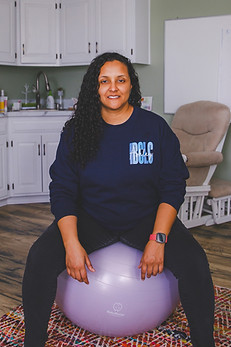 Ashley Haden-Peaches, Doula and IBCLC, wearing a navy sweatshirt and black pants, sitting on a birth ball, in her office.