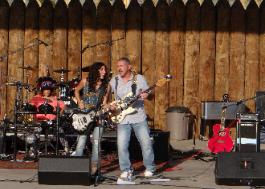 Two musicians with guitars singing together, with a drummer in the back