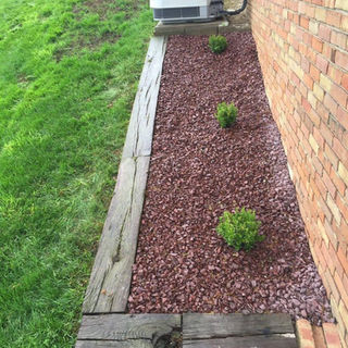 Red mulch landscaping with three small shrubs and a wooden border beside a brick house