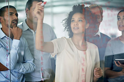 Image of a woman writing on a glass wall surrounded my four colleagues looking at her work. This is listed under 3PV's "Research for Policy Action" service offering.