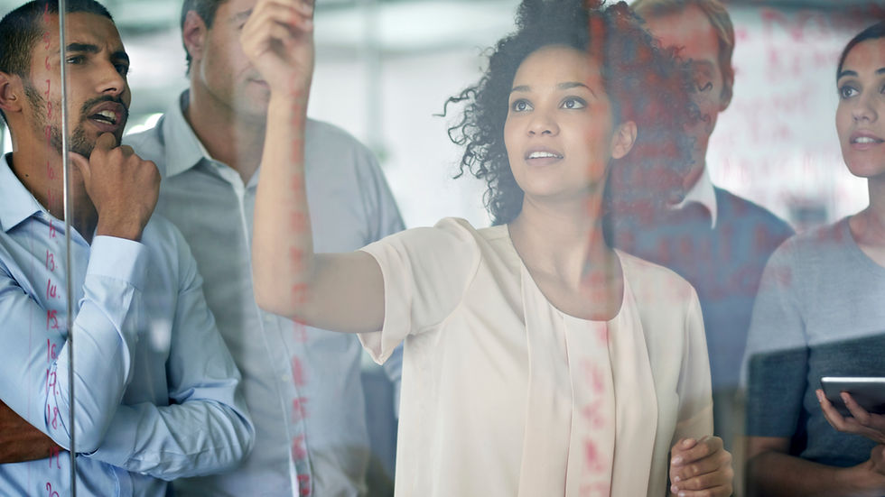 Four people brainstorm, one writing on glass with red marker, others observing. Office setting, casual attire, focused expressions.