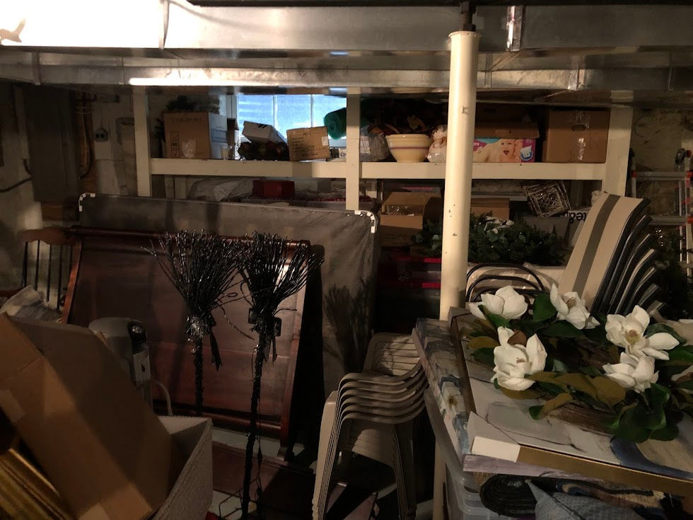 a before photo of a basement Organization Pending decluttered and organized with a client - piles of stuff and boxes with furniture pieces are on the floor, with cluttered shelves in the back