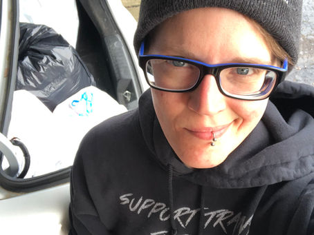 Owner/Operator of Organization Pending, Tabi (they/them), stands smiling in front of their car's open trunk, full of donation bags. They are wearing black rimmed glasses, a hat with the company's logo and name on it, and a hoodie that says "Support Trans Futures".