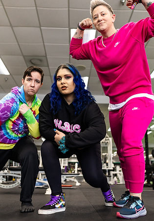 three people are posing in classic muscle poses, the two on the left are crouching. the person on the left has a tie die hoodie, black jeans, and short hair, the person in the middle has long blue hair, and is wearing a black hoodie and pants, and the person on the right has short blonde spiky hair, wearing matching pink pants and sweater