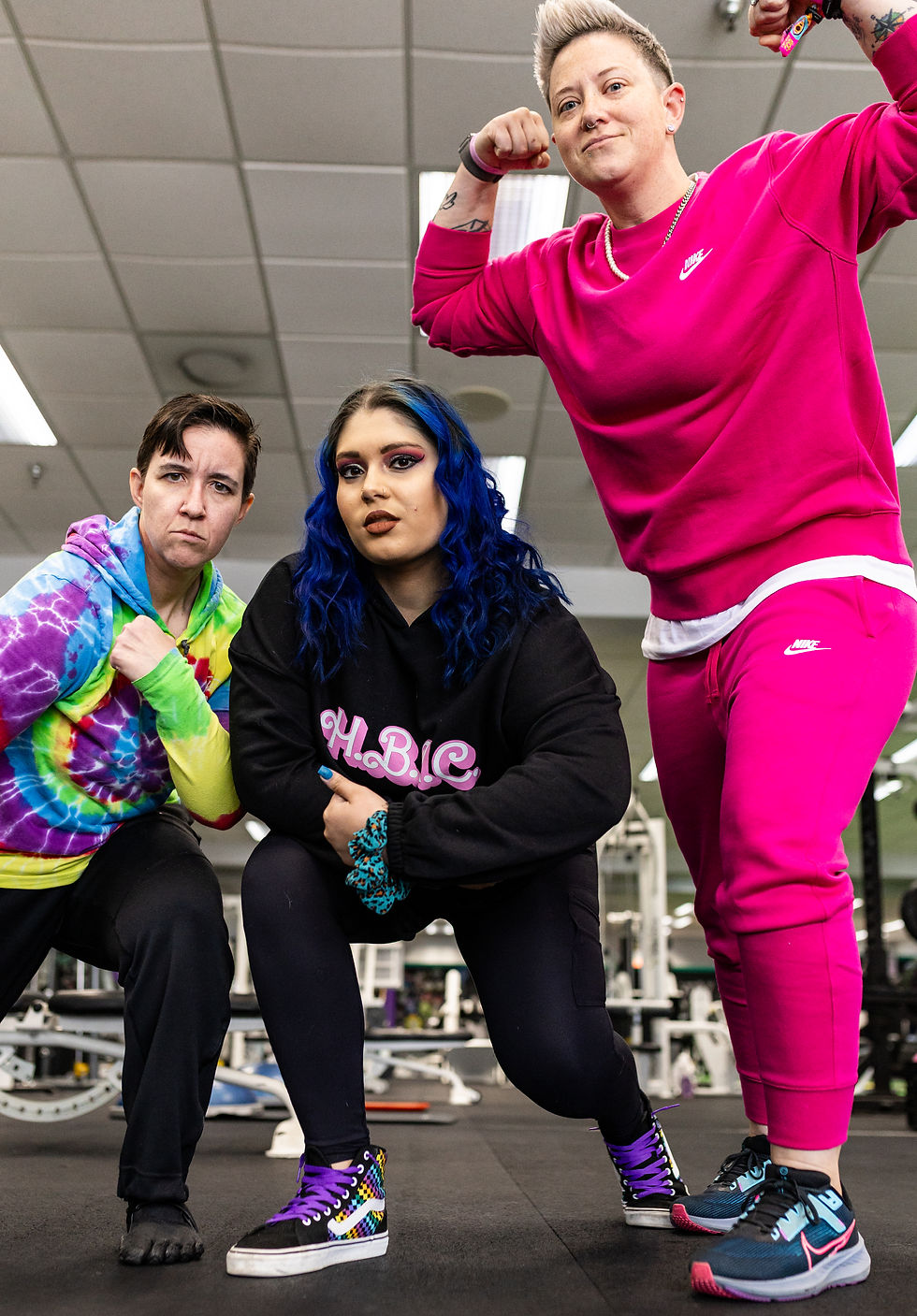 three people are posing in classic muscle poses, the two on the left are crouching. the person on the left has a tie die hoodie, black jeans, and short hair, the person in the middle has long blue hair, and is wearing a black hoodie and pants, and the person on the right has short blonde spiky hair, wearing matching pink pants and sweater