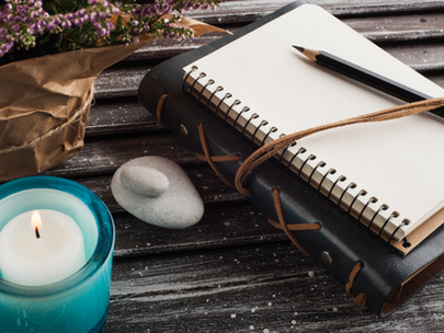 Journal with a pencil, lit candle, a bunch of dry pink flowers and a couple of stones. All displayed over a desk.