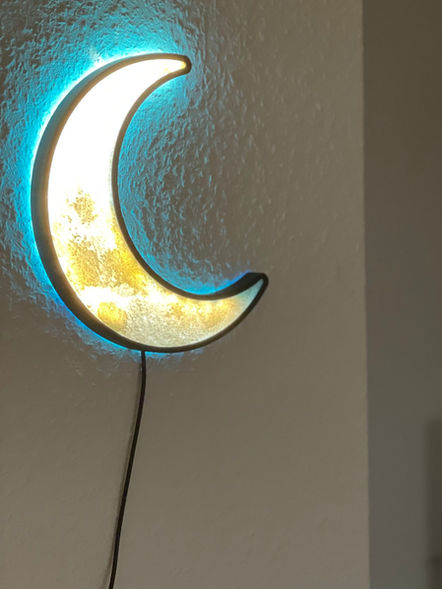 Eine erleuchtete Mondsichel mit blauem Schein hängt an einer Wand. Im Hintergrund ist eine Kommode mit Pflanze und Kerzenständer zu sehen.