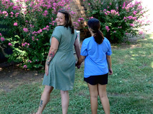 A woman and her daughter walk toward some flowers away from the camera. The woman looks over her shoulder and smiles. She wears a green striped dress and has long brown hair. Her daughter wears a blue t-shirt and black shorts and has long brown hair.