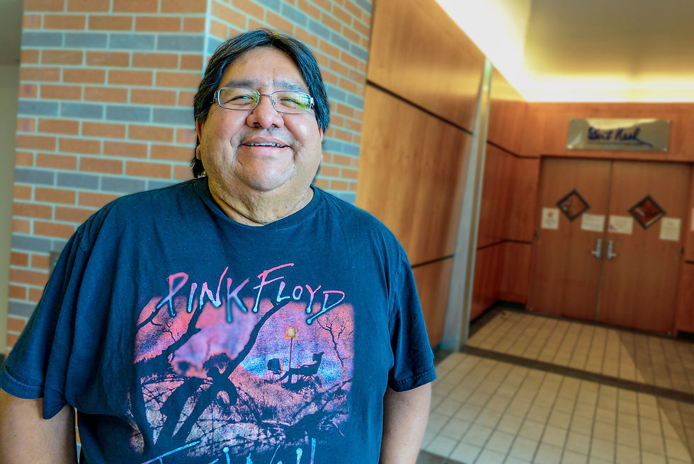 A man smiles in front of a brick wall. He wears glasses and a Pink Floyd t-shirt.