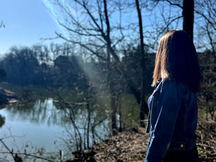 A woman looks out on a pond in a wooded area.
