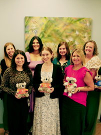A group of leaders from DCCCA and Bert Nash Center smile in front of a painting of a dandelion. Some hold stuffed koala bears with Mental Health First Aid shirts on.