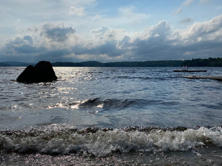A glossy lake peppered with rocks and a nearby dock as sun shines down on the surface