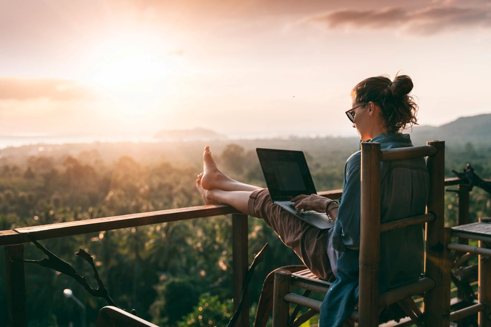 A women sitting comfortably on her deck, facing the sunset, working on her laptop