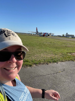 woman in hat with sunglasses and blue top, pavement and grass with a airplane in the background