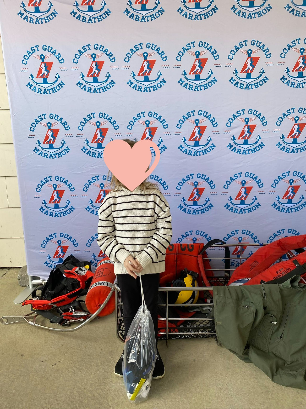 Coast Guard Marathon logo on back drop, rescue swimmer basket and search and rescue gear behind little girl with race packet.  Little girl in dark pants and long sleeve white sweater with black stripes.