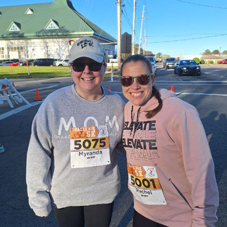 white building with green roof in background, two women on a street, one wearing a hat, sunglasses, gray sweatshirt and black pants, the other wearing sunglasses, a pink sweatshirt and black pants.