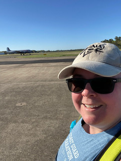 woman in hat with sunglasses and blue top, pavement and grass with a airplane in the background