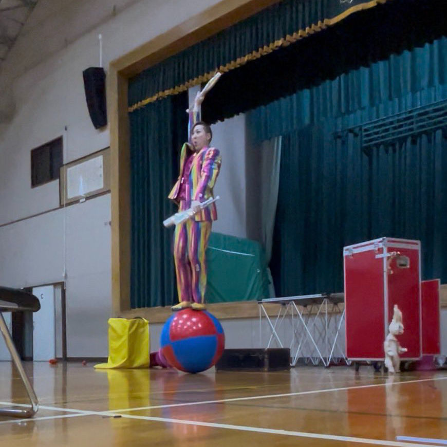北海道 ピエロ・TeTe(テテ)が小学校の体育館ステージでバランスボールの上に立ち、ジャグリングを披露しているパフォーマンスの様子。子ども向けイベントで会場を盛り上げる大道芸シーン