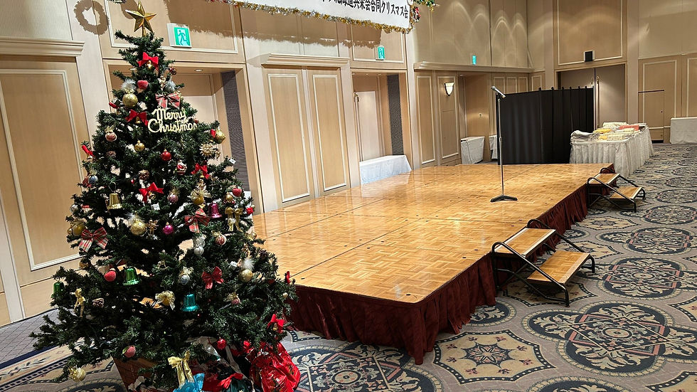 ホテル宴会場に設置されたクリスマスツリーとステージ。企業のクリスマス会のために準備されたマイクや装飾が並び、温かい雰囲気の会場が整っている様子