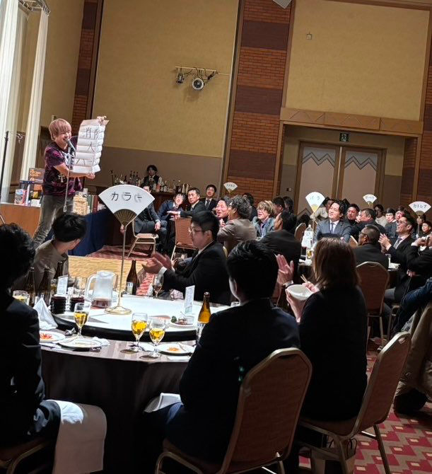 帯広市の企業忘年会の宴会場で、北海道のプロマジシャン・アッキーが予言マジックを披露している様子。紙に書かれた予言を観客に見せ、テーブル席の参加者たちが驚きと拍手で盛り上がっている場面