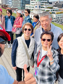 Group selfie of smiling tourists on a hillside overlook, with city buildings and harbor behind; two women flash peace signs.