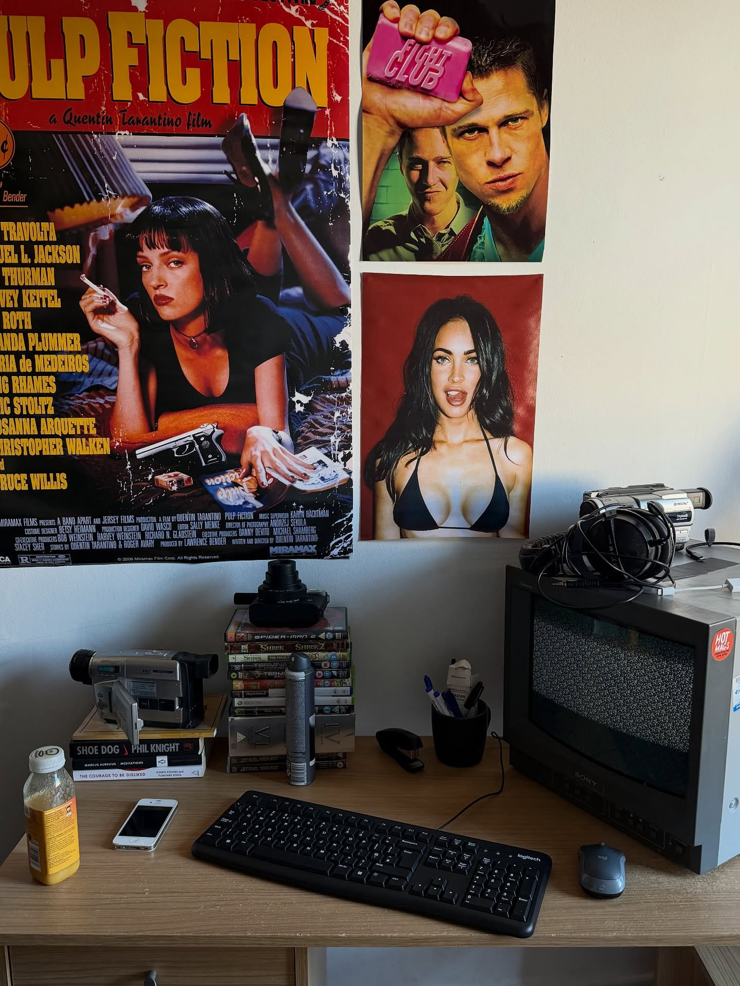 Desk setup in a bedroom or studio with a keyboard, mouse, phone, camcorder, VHS tapes, and a small CRT television. Above the desk, several bold movie posters cover the wall, creating a retro, film-inspired workspace with vintage media equipment and everyday items.