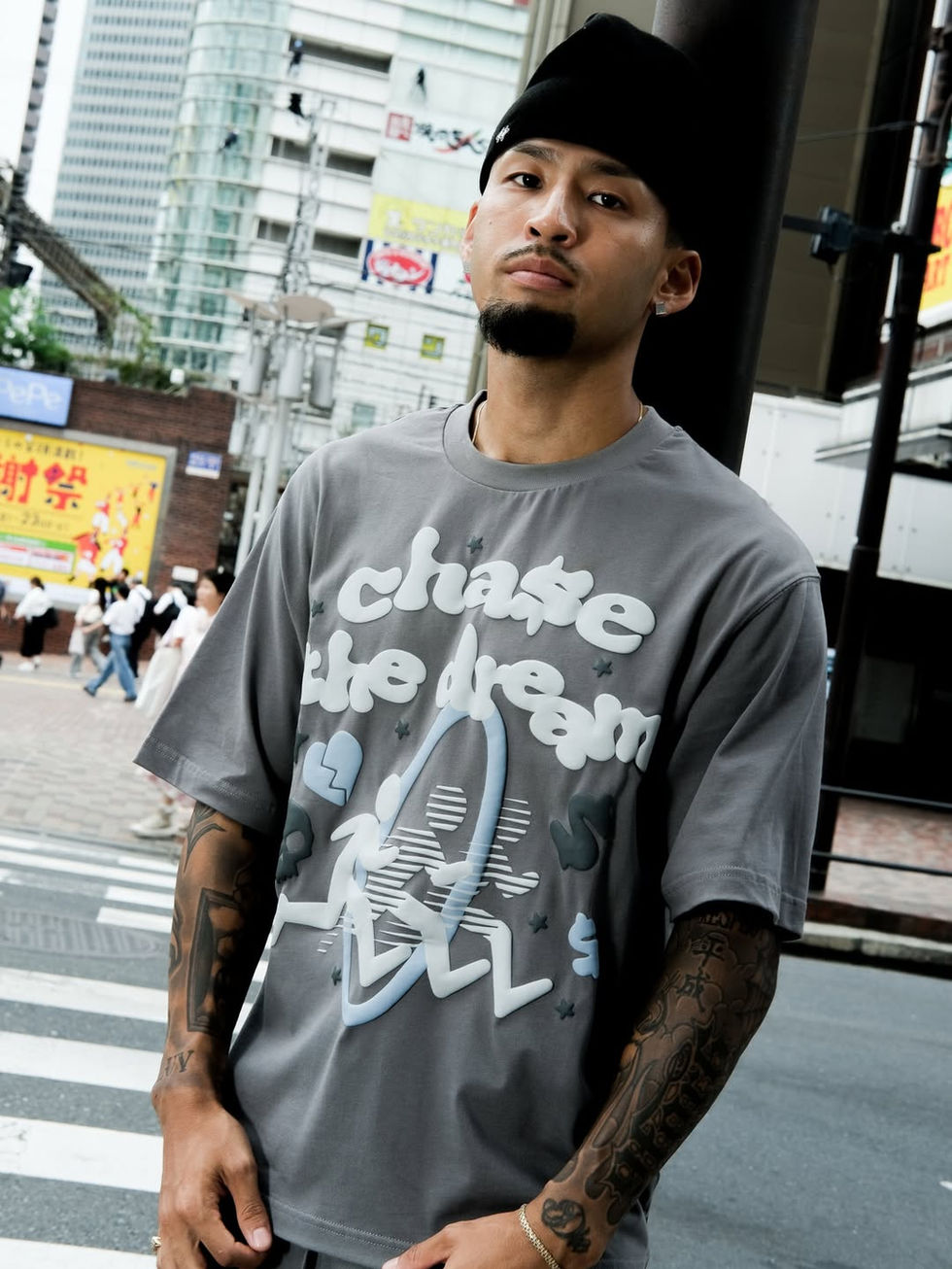Person wearing a grey graphic T-shirt and black beanie stands on a city street. The shirt features large stylised text and illustration, while tall buildings, street signs, and pedestrians appear blurred in the background, creating an urban daytime setting.