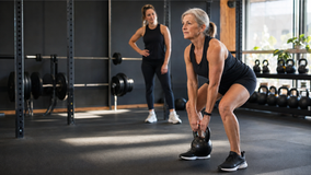 Older woman in black workout clothes lifts a kettlebell in a gym while a younger woman watches nearby.