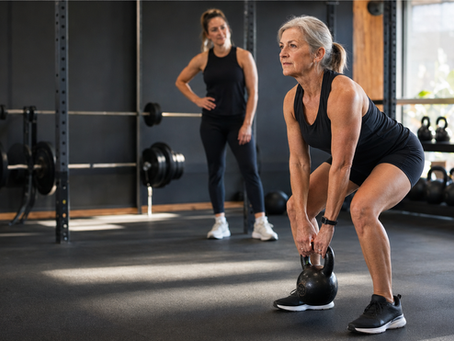 Older woman in black workout clothes lifts a kettlebell in a gym while a younger woman watches nearby.