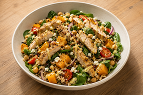 Bowl of grilled chicken salad with spinach, chickpeas, cherry tomatoes, squash, red onion, and creamy dressing on a wooden table