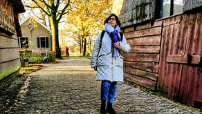 אישה במעיל אפור וצעיף סגול הולכת על שביל מרוצף באבנים, מרופד בעצי סתיו ובנייני עץ, תחת אור שמש חמים.