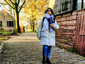 אישה במעיל אפור וצעיף סגול הולכת על שביל מרוצף באבנים, מרופד בעצי סתיו ובנייני עץ, תחת אור שמש חמים.