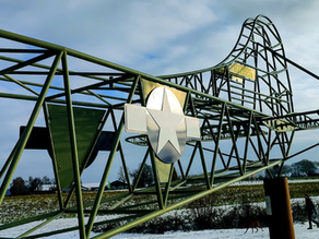 פסל דאון מתכת ירוק עם סמל כוכב על שדה מושלג. שמיים מעוננים, אדם הולך עם כלב ברקע.