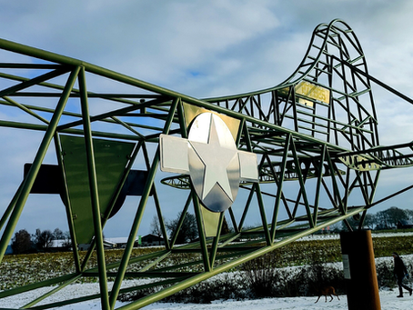 פסל דאון מתכת ירוק עם סמל כוכב על שדה מושלג. שמיים מעוננים, אדם הולך עם כלב ברקע.