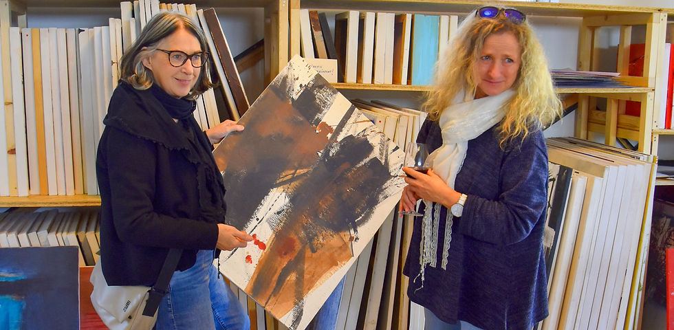 Zwei Frauen halten ein abstraktes Gemälde mit dunklen Farben. Regale voller Leinwände im Hintergrund. Eine Frau hält ein Weinglas.