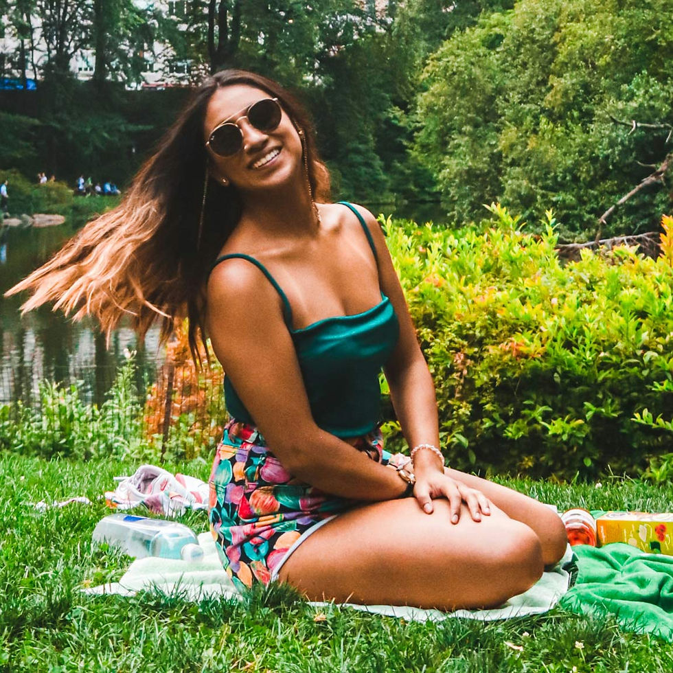 Woman in sunglasses sits on grass, wearing a green top and colorful shorts. She's smiling, surrounded by greenery near a pond.