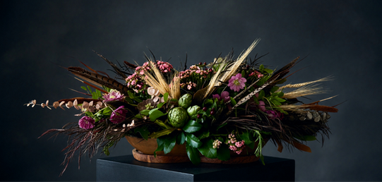 Low elongated autumn centerpiece with artichokes, wheat, feathers, pink flowers, berries, dried foliage, and glossy greenery in wooden vessels.