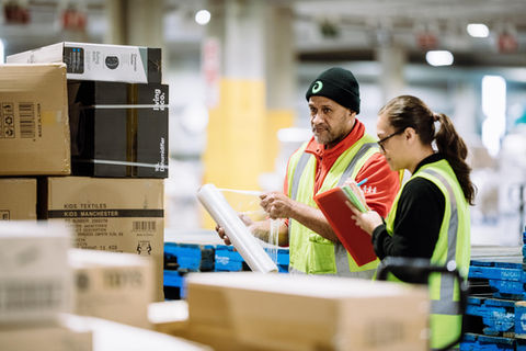 Two warehouse workers reviewing documents surrounded by packages and boxes at work.