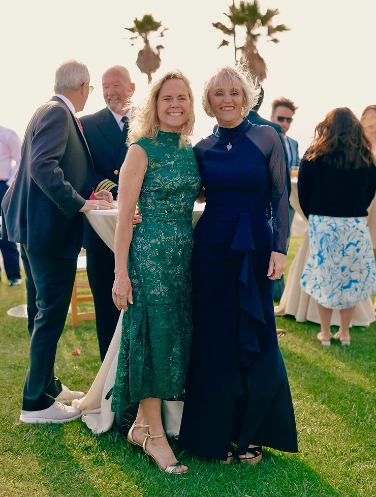 Two smiling women in fancy dresses, standing, with people smiling in the background.