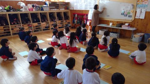先生がホワイトボードに何かを書いている。園児は床に座って授業を受けています。