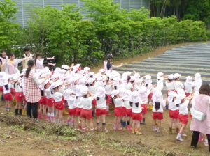 幼稚園児が体操服姿で手遊び。周囲には緑の木々、階段があります。おたのしみ会。
