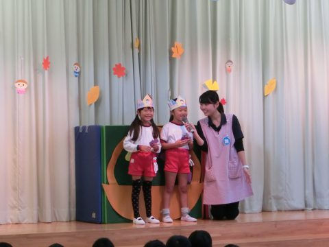 二人の女の子と先生がステージで歌っています。可愛らしい衣装を着ています。年中組　園外保育