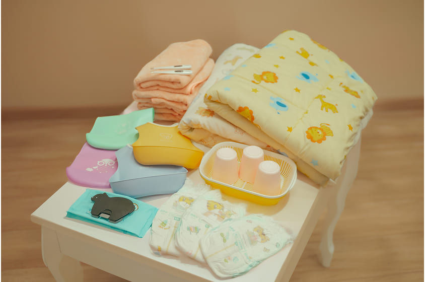 ベビー用品がテーブルに並んでいる様子。タオル、スタイ、おむつなど。マジ