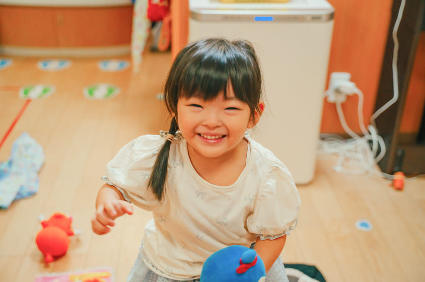 笑顔の女の子、白いシャツを着て楽しそうに微笑む、周りにぬいぐるみが、マジオブルームキッズ