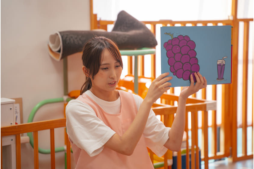 保育士が絵本を見せる、ブドウの絵。先生の1日 幼稚園の教室で、子供達へ読み聞かせ